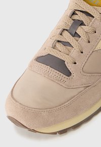Sneaker beige con una combinazione di texture in suede e tessuto, lacci tondi e punta rinforzata; accenti grigi sottili e dettagli di cucitura.