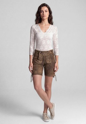 Frau steht und trägt ein weißes Spitzen-Longsleeve-Top, braune bestickte Ledershorts und beige Sneaker vor einem schlichten grauen Hintergrund.