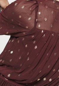 Maroon topp i lätt, genomskinlig tyg med spridda metalliska guldprickar, med samlade detaljer och flödande fall.