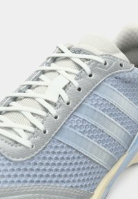 Αθλητικό παπούτσι ανοιχτού μπλε χρώματος με επάνω μέρος από δίχτυ, λευκά κορδόνια και τρεις εμφανείς γκρι ρίγες. Διαθέτει υφασμένο σχέδιο και μαλακή, μαξιλαρωτή σόλα.