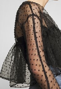 Top nero trasparente con maniche a sbuffo, texture a pois e design a strati. Il materiale è leggero e leggermente trasparente.