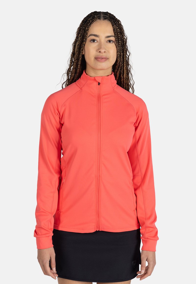 Ljus korallfärgad zip-up-jacka gjord av elastiskt tyg, med hög krage och långa ärmar, i kombination med en svart kjol.