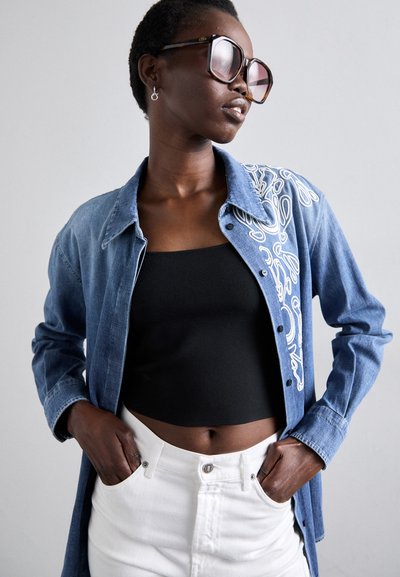 Camicia di denim con ricami bianchi sulla spalla, indossata sopra una canotta nera, abbinata a pantaloni bianchi a vita alta. Grandi occhiali da sole scuri.