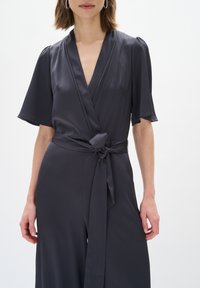 Jumpsuit grigio avvolgente con maniche corte, tessuto testurizzato, scollo a V profondo, vita con cintura e gambe ampie e fluttuanti.