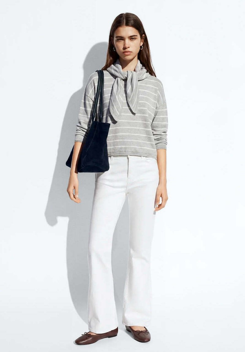 Femme portant un pull rayé gris, pantalon blanc, ballerines marron, portant un sac cabas noir, debout devant un fond blanc avec une ombre.