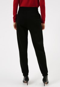 Zwarte sweatpants met een relaxed fit, elastische tailleband en geribbelde boorden, gecombineerd met zwarte enkellaarzen met hak. Achteraanzicht.