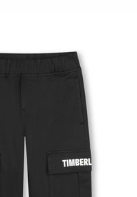 Svarte cargo-bukser laget av et glatt stoff, med en elastisk midje, sidelommer og en hvit "TIMBERLAND"-logo på cargo-lommen.
