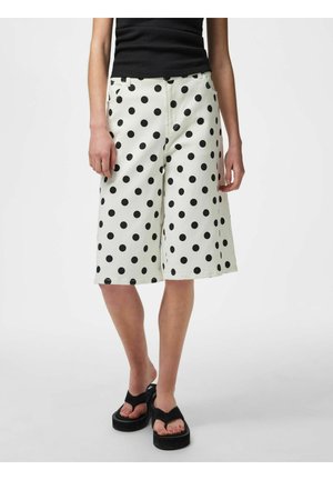 Persona che indossa pantaloncini bianchi al ginocchio con pois neri, top nero senza maniche e sandali platform neri su sfondo semplice.
