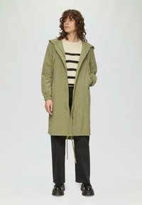 Olivenfarbenen langen Mantel mit Kapuze, Kordelzug in der Taille und Reißverschluss. Gestreifter beigefarbener und schwarzer Pullover darunter. Schwarze Hose und Schuhe.