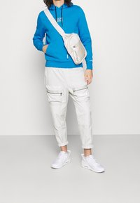 Sudadera azul con logo negro, pantalones cargo blancos con bolsillos con cremallera y zapatillas blancas. Se lleva una bandolera de color beige claro.