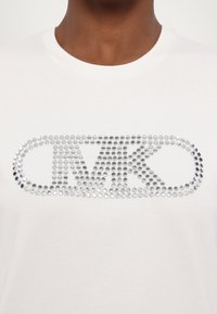 White cotton t-shirt featuring a large silver rhinestone logo "MK" in the center, with a rounded rectangular outline. Soft texture.