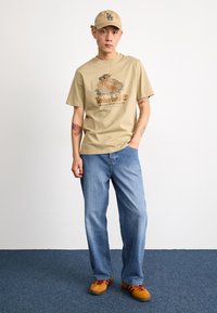 T-shirt beige à manches courtes avec un logo Timberland imprimé, associé à un jean bleu clair et des baskets orange avec des accents bordeaux.