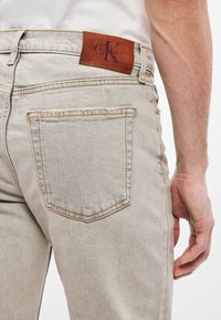 Gaiši pelēkas džinsa bikses ar ādas Calvin Klein logotipu uz jostas vietas, redzama aizmugurējā kabata un smalkas šuvju detaļas.