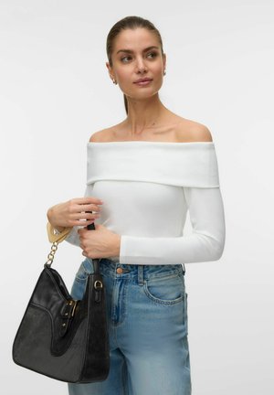 Vrouw met paardenstaart die een witte off-shoulder top met lange mouwen en blauwe jeans draagt, en een zwarte handtas met gouden accenten vasthoudt.