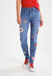 Blå denimjeans med slim fit, dekorerade med färgglada broderade lappar i olika mönster på båda benen. Vita skor med röda detaljer.