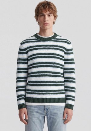 Jeune homme aux cheveux bouclés portant un pull tricoté rayé vert et blanc et un jean bleu clair, debout devant un fond uni.