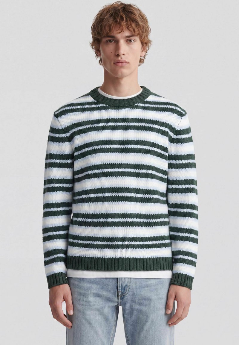 Jeune homme aux cheveux bouclés portant un pull tricoté rayé vert et blanc et un jean bleu clair, debout devant un fond uni.
