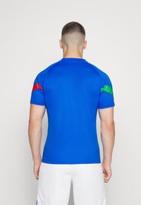 Niebieska koszulka sportowa z czerwonymi i zielonymi akcentami na rękawach, wykonana z fakturowanej tkaniny i z zaokrąglonym dekoltem. Pokazano widok z tyłu.