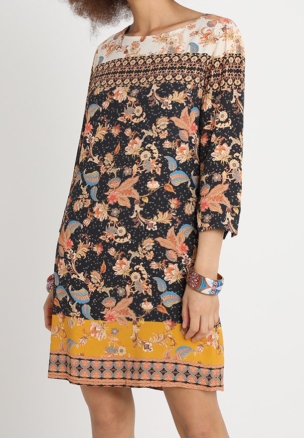 Femme portant une robe mi-longue avec des motifs floraux en noir, blanc et jaune, et un bracelet coloré à motifs au poignet.