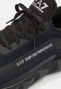 Black sneaker featuring a textured mesh upper, flat laces, and a rubber sole with a geometric pattern; logo displayed on the side.