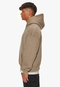 Beige Kapuzenpullover mit einer Vordertasche, oversized Design und gerippten Bündchen. Das Material wirkt weich mit einer glatten Textur.