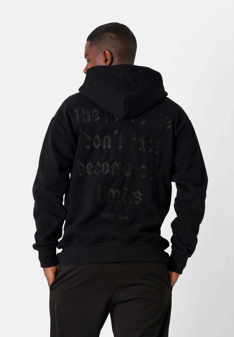 Schwarzer Hoodie mit verstellbarem Kapuzenzug. Auf der Rückseite befindet sich geprägter Text in einer kontrastierenden dunklen Farbe. Rippstrickbündchen und Saum. Glatte Textur.