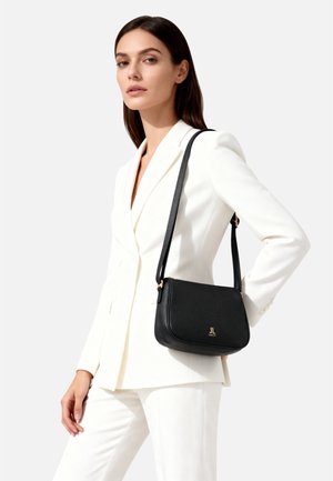 Zwarte schoudertas van getextureerd leer met een gebogen vorm, goudkleurig logo en verstelbare band, gedragen door een model in een wit pak.