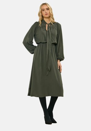 Vrouw met lang blond haar, gekleed in een midijurk met lange mouwen in olijfgroen met een nekstrik, zwarte panty's en zwarte hakken, staand met één hand op haar heup.