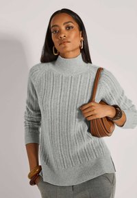 Eine Frau trägt einen hellgrauen, gerippten Rollkragenpullover, graue Hose, goldene Creolen, braune Armbänder und hält eine braune Leder-Schultertasche.