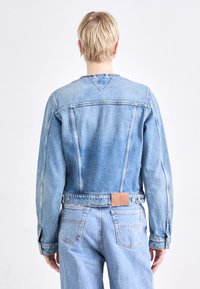 Chaqueta de mezclilla azul claro con diseño entallado, costuras visibles y una etiqueta de cuero con la inscripción "TOMMY JEANS" en la parte trasera. Mangas largas.