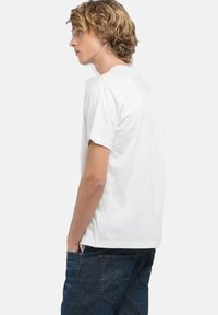 Witte t-shirt met ronde hals, gemaakt van katoen, korte mouwen, relaxte pasvorm, gedragen met donkere spijkerbroek, gezien vanaf de zijkant.