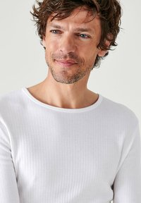 Witte geribbelde lange mouwen shirt met een ronde halslijn. De stof heeft een gladde textuur en een getailleerde pasvorm. Subtiele detailing voegt diepte toe.