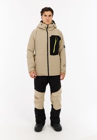 Beige isolierte Jacke mit einer schwarzen Brusttasche und einem gelben Reißverschluss, kombiniert mit schwarzen und beigen Hosen. Verfügt über eine Kapuze und eine verstellbare Passform.