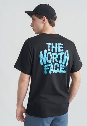 Giovane uomo con berretto nero indossa una maglietta nera con grande scritta blu "THE NORTH FACE" sulla schiena, in piedi contro uno sfondo chiaro e uniforme.