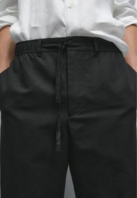 Pantaloni neri con vita elastica e cordino, indossati con una camicia bianca a maniche lunghe, mani nelle tasche.