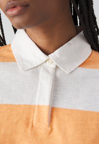 Polohemd mit horizontalen Streifen in sanftem Orange, Weiß und Hellgrau. Es hat einen weißen Kragen und eine Knopfleiste. Die Textur wirkt leicht.