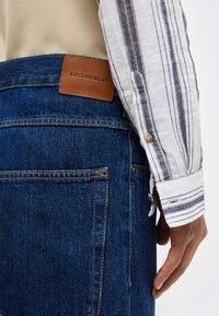 Άτομο που φοράει μπλε τζιν από denim με καφέ ετικέτα LINDEBERGH και λευκό ριγωτό μακρυμάτικο πουκάμισο, το χέρι ακουμπά δίπλα στο τζιν.