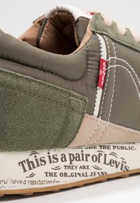 Baskets vert olive avec matériaux mixtes : tissu lisse et daim. Coutures blanches remarquables, étiquette rouge Levi's et texte imprimé sur la semelle.