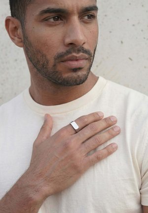Man with short beard and neutral expression wearing white shirt, hand with silver ring resting on his chest against light background.