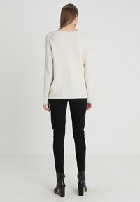 Světle béžový svetr s texturovaným pletením, zdobený cvočky na ramenou, kombinovaný s černými skinny kalhotami a černými kotníkovými botami na vysokém podpatku.
