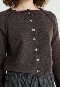 Cardigã castanho texturizado com um decote redondo e cinco botões à frente, apresentando uma bainha canelada e mangas raglan. Saia preta com riscas verticais visível por baixo.