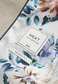 Etiqueta de características "PRÓXIMA CONFECCIÓN" con tamaño "40R" en tela con estampado floral; la etiqueta dice "AJUSTE DELGADO" en un diseño simple y plano.