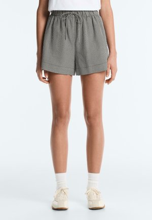 Žena v čierno-bielych károvaných krátkych nohaviciach s elastickým pásom s povrazcom, bielych ponožkách a krémových teniskách na bielom pozadí.