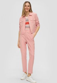 Lichtroze denimjack met twee voorzakken, gedragen over een multikleurig gestreept shirt. Bijpassende roze broek en witte sneakers maken de look af.