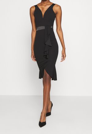 Vrouw draagt een nauwsluitende zwarte mouwloze jurk met ruches aan de voorzijde en zoom, gecombineerd met zwarte hooggehakte schoenen.