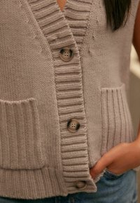 Jasnoszara dzianinowa kamizelka z prążkowanym kołnierzem i obszyciem, z trzema okrągłymi guzikami i dwoma przednimi kieszeniami. Miękka tekstura i luźny krój.