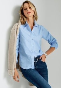Hellblau gestreiftes Hemd mit Button-Down-Kragen, kombiniert mit einem beigefarbenen gerippten Cardigan. Das Model trägt dunkle Jeans und silbernen Schmuck.