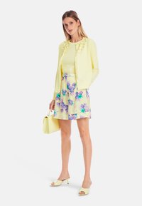 Yellow cardigan with decorative buttons, yellow top, floral-patterned skirt in light yellow with purple flowers, yellow handbag, and shoes.