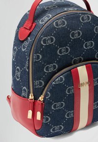 Zaino in denim con accenti in pelle rossa, zip dorata e dettagli a righe. Decorato con logo su una superficie blu texturizzata.