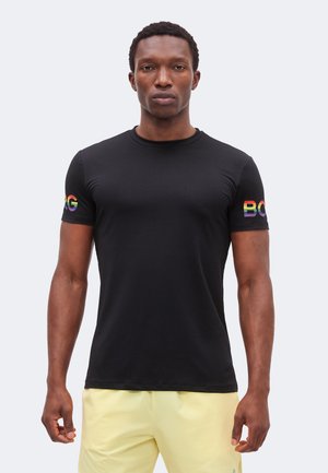 Mężczyzna stojący przodem, ubrany w czarną koszulkę z napisem "BOSS" w kolorach tęczy na rękawach oraz jasnżółte szorty.